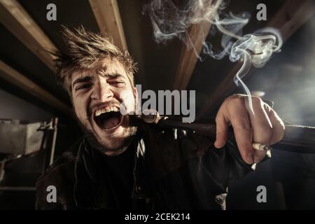 Young beard guy with opened mouth with burning cigarette and riffle in hand on garret Stock Photo
