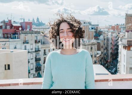 Pretty young brunette with curly hair smiling white standing on background of beautiful city. Stock Photo