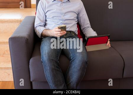 The man uses his mobile phone sitting on the sofa and in the meantime looks at the tablet, intensive use of technology Stock Photo