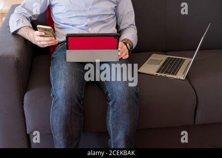 The man uses his mobile phone sitting on the sofa and in the meantime looks at the tablet and laptop, intensive use of technology Stock Photo