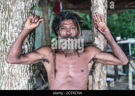 African American bearded male with dreadlocks and upped hands smoking cigar near trees Stock Photo