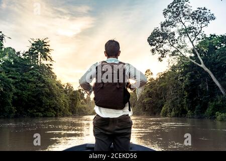 Back view young guy with knapsack standing on bow of ship sailing on river between exotic forest on shores in Malaysia Stock Photo