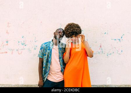 Cheerful African-American man and Woman in stylish outfits smiling and looking at camera while standing near concrete wall on street Stock Photo