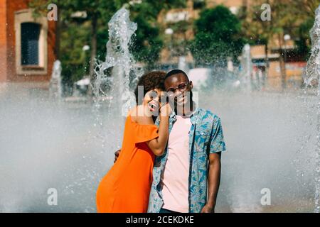 Handsome African-American man hugging pretty Woman and laughing while standing near fountain together Stock Photo