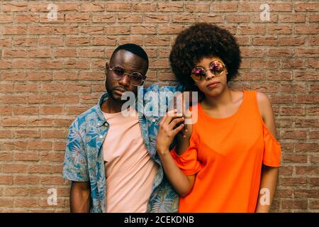 Attractive African-American man and Woman in stylish outfits laughing and hugging while standing near brick wall on street Stock Photo