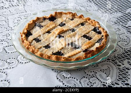 Fresh hot blueberry pie in a glass pie pan on a lace tablecloth.  Steam coming off this lattice crust homemade dessert. Stock Photo