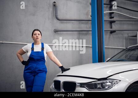Woman in blue overalls leans on the hood of the car and poses Stock Photo
