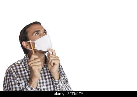 one young adult latin or hispanic white man wearing t-shirt and face mask holding pencil in hand thinking of a project on white background Stock Photo