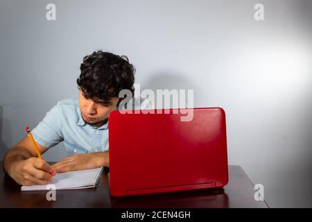 Young hispanic wavy hair man studying from home with a laptop and writing with a pencil in a notebook Stock Photo