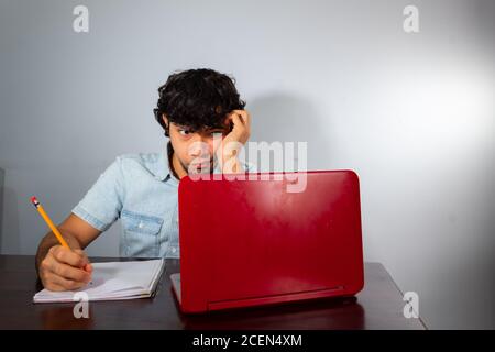 Young hispanic wavy hair man studying from home with a laptop and writing with a pencil in a notebook Stock Photo