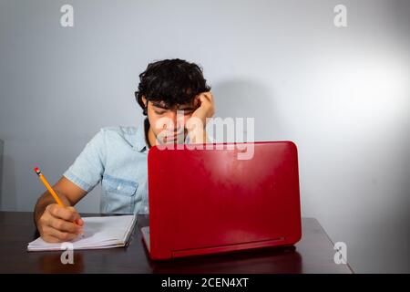 Young hispanic wavy hair man studying from home with a laptop and writing with a pencil in a notebook Stock Photo
