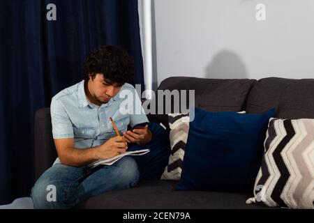 Young hispanic man with wavy hair studying from home with a cellphone Stock Photo