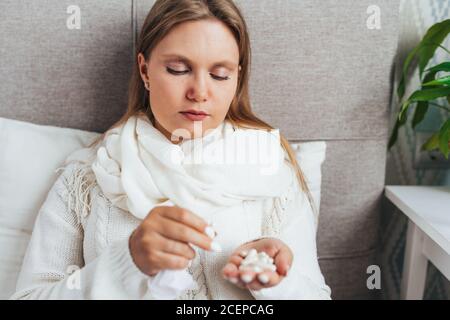 Young woman with a handful of pills lies in bed at home wearing sweater and a warm scarf. Flu, cold, sickness Stock Photo