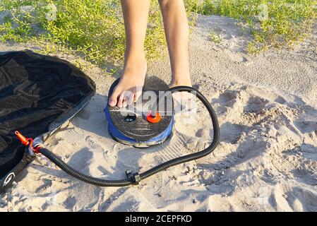 A woman with air foot pump pumps an inflatable mattress or air bed at sandy beach. Foot inflates air mattress with foot pump on sand. Stock Photo