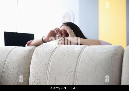 Woman looks out from behind sofa and holds her hands through binoculars. Stock Photo