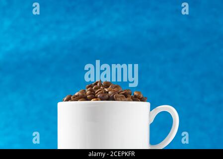 International day of coffee concept. close-up white coffee cup full of coffee beans on blue background. Stock Photo