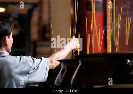 a man places burning incense sticks at an altar in an old Chinese Buddhist Temple in Southeast Asia Stock Photo