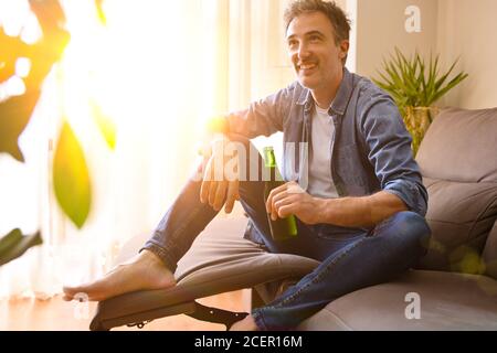 Relaxed smiling man drinking a beer resting sitting on a sofa in the living room at home. Stock Photo