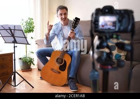 Video Blogger teaching to play the guitar with a camera recording in his living room Stock Photo