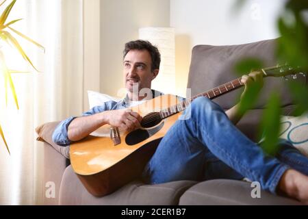 Relaxed and smiling man lying on a sofa playing the guitar in a room at home Stock Photo