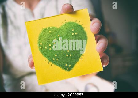 Woman's hands holding sticky note with green heart with yellow flowers, natural and ecological life style concept, gardening in pure, healthy environm Stock Photo