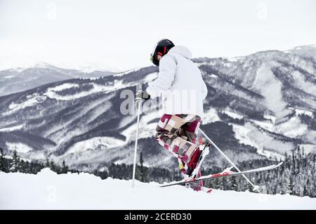 Back view of alpine skier in jacket and helmet skiing in mountains. Man freerider with ski sticks walking through the snow with beautiful snowy hills on background. Concept of winter sport activities. Stock Photo