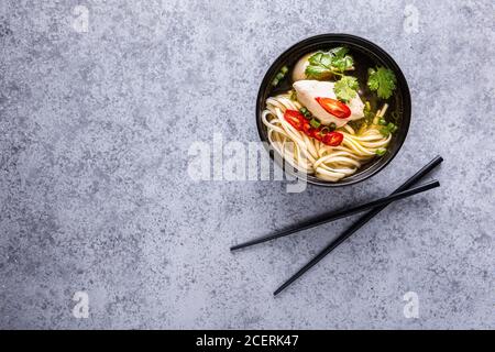 Spicy asian soup. Chicken bouillon and meat, red chilli pepper, cilantro, scallions and japanese noodles in black bowl with chopsticks on gray surface Stock Photo