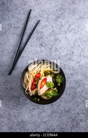 Spicy asian soup. Chicken bouillon and meat, red chilli pepper, fresh cilantro and scallions, japanese noodles in black soup bowl with chopsticks on g Stock Photo
