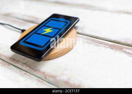 Concept of modern technologies. On the wireless charger is a black smartphone with a blue battery icon. White wooden background with diagonal stripes. Stock Photo