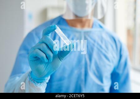 Horizontal medium close-up shot of unrecognizable medical worker wearing protective uniform holding ampoule with transparent liquid Stock Photo