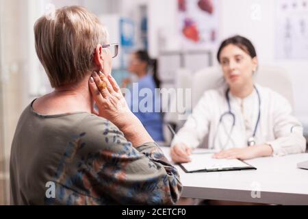 Mature woman suffering neck pain during consultation with doctor in hospital cabinet. Senior patient examination. Stock Photo