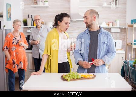 Man proposing wife during family lunch. Shocked young woman looking at engagement ring while holding a glass of wine Stock Photo