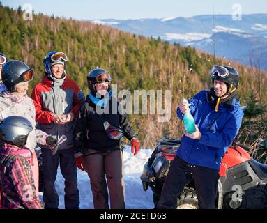 Joyful young man opening champagne while friends looking at him and smiling. Cheerful people celebrating successful trip in winter mountains. Concept of celebration, active leisure and quad biking. Stock Photo