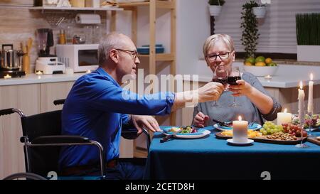 Senior old man in wheelchair dining with his cheerful wife sitting at the table in the kitchen. Imobilized paralyzed handicapped elderly husband having romantic dinner Stock Photo
