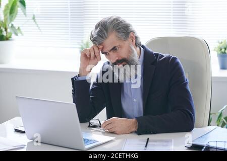 Portrait of concentrated mature male office worker wearing eyeglasses sitting at office desk and reading information on laptop Stock Photo