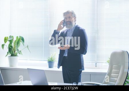 Horizontal medium long shot of mature businessman with beard on face and stylish haircut standing in office room talking on phone Stock Photo