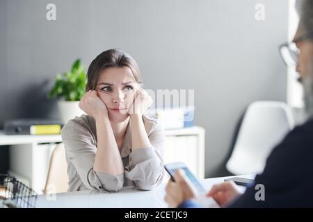 Over-the-shoulder portrait of sad young woman feeling anxiety sitting in front of psychologist looking at him Stock Photo