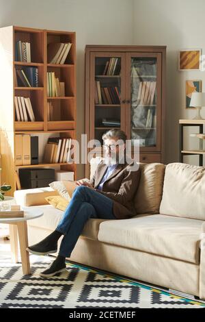 Vertical shot of handsome bearded man in stylish outfit sitting relaxed on sofa in modern office room making notes Stock Photo