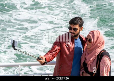 a young asian or ethnic bame couple on holiday taking a photgraph with a mobile phone device using a selfie stick on the isle of wight ferry. Stock Photo