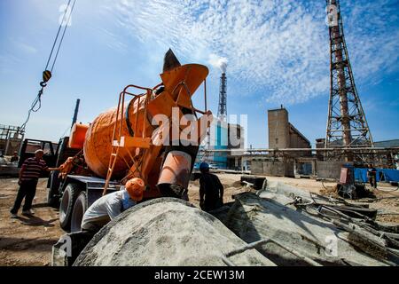 Old phosphate fertilizer plant in modernization. Mobile crane, workers, orange mixer truck, cement buckets, industrial building, factory chimneys on b Stock Photo