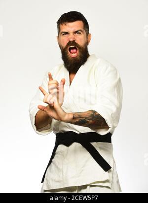 Man with beard in white kimono on white background. Karate man with mad excited face in uniform. Healthy lifestyle and sports concept. Aikido master with black belt practices attack or defense posture Stock Photo