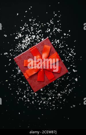 Red box with a gift on a dark background with many scattered sparkles or confetti nearby. Festive concept. Stock Photo