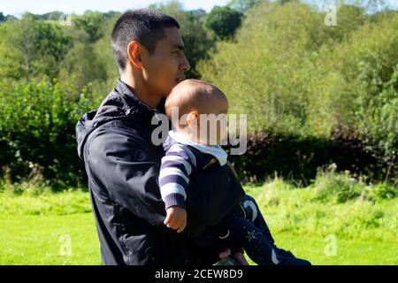 Asian man 30s profile side view holding child baby son 6 months wearing anorak in the countryside Wales UK  KATHY DEWITT Stock Photo