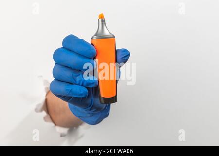 Hand in a latex glove holding an orange highlighter pen through torn white wall Stock Photo