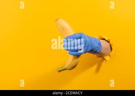 Hand in a latex glove holding a banana through torn yellow wall Stock Photo