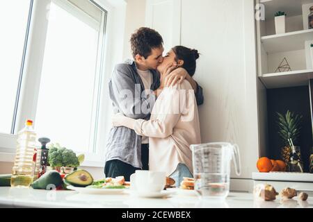 Caucasian couple kissing in the kitchen while preparing food near the window Stock Photo