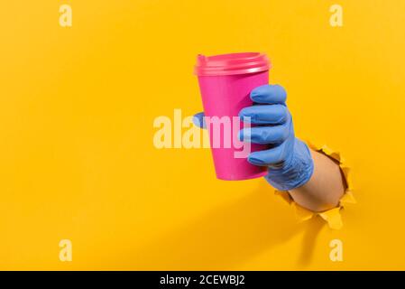 Hand in a latex glove giving a pink coffee cup through torn yellow wall Stock Photo