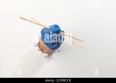 Hand in a latex glove holding wooden chopsticks through torn white wall Stock Photo