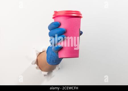 Hand in a latex glove holding a pink cup of cofee through torn white wall Stock Photo