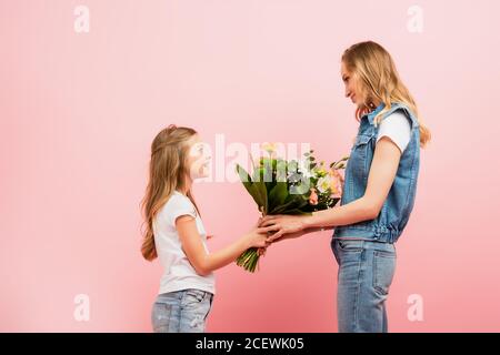 side view of girl presenting bouquet of flowers to mother wearing denim clothes isolated on pink Stock Photo